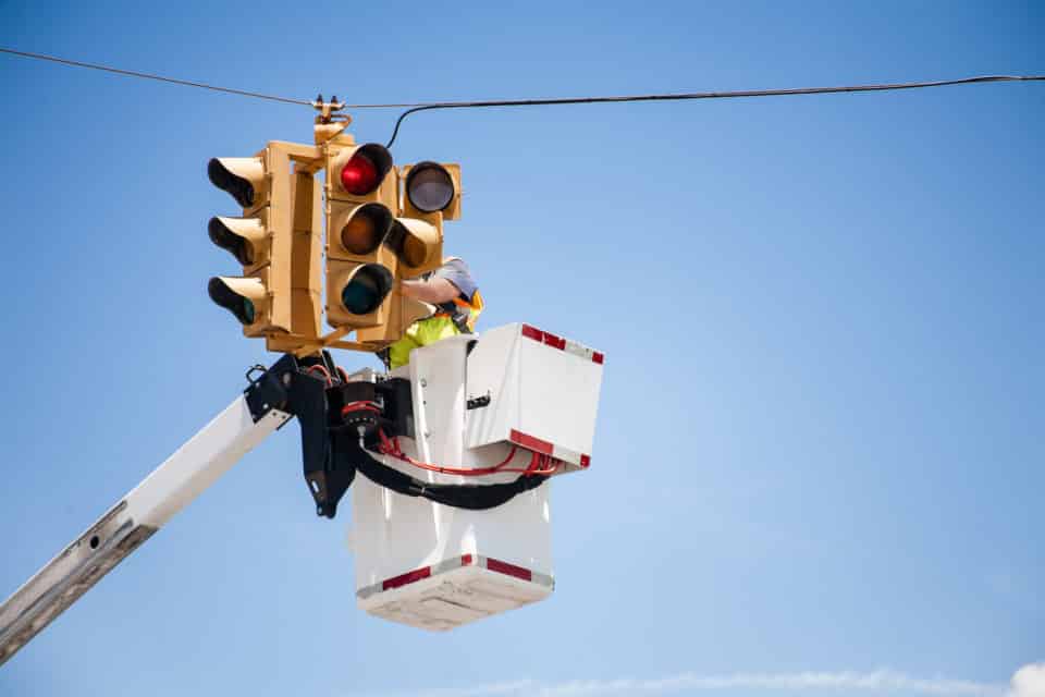 Unrecognizable utilities worker repairs a traffic signal, light using a bucket lift at a road intersection in USA. Clear blue sky. One man wearing safety vest.